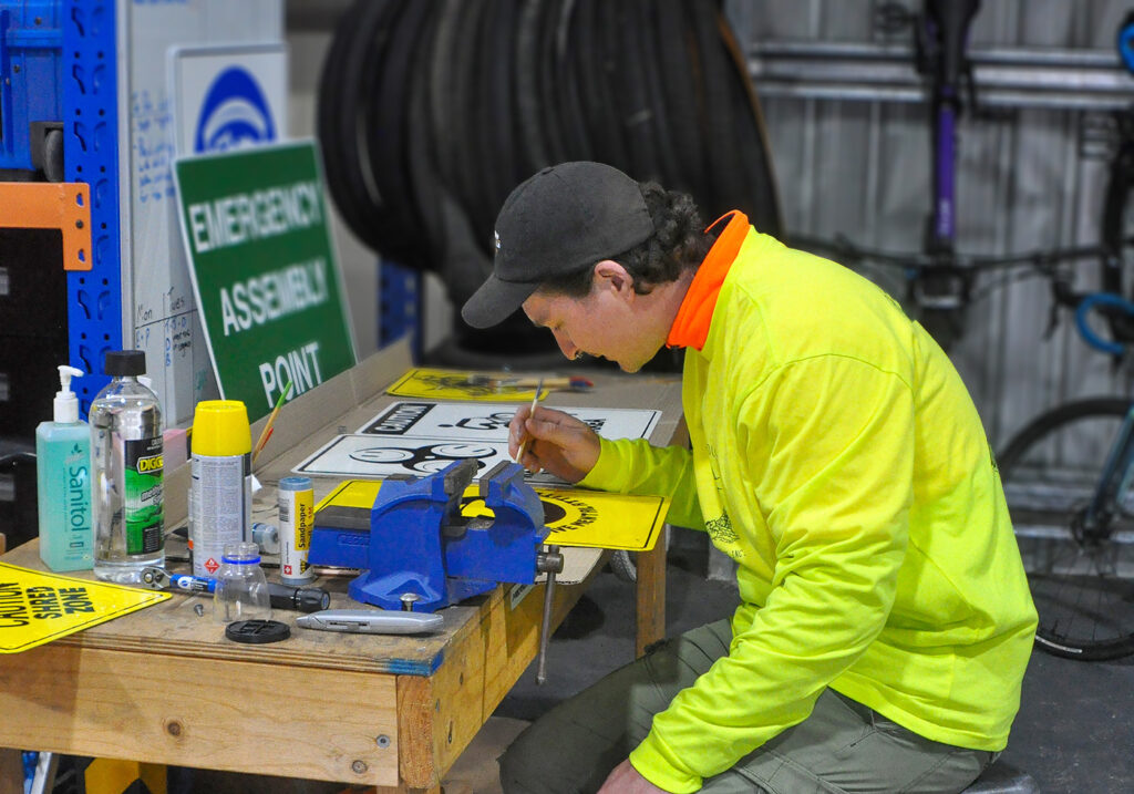 Daniel is seated at a timber table inside the Lighthouse Youth Projects building and is creating a yellow sign with a positive message for young people visiting the building. Daniel wears a black cap and a fluorescent yellow jacket with a bright orange collar.  