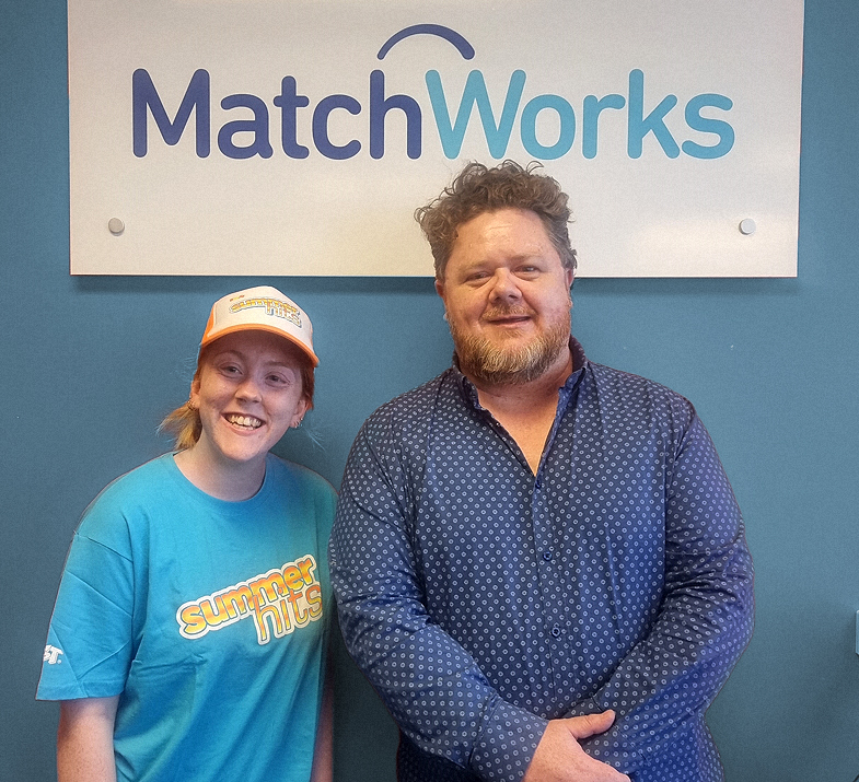 Tiahnee stands next to Nick from Oaklands Park MatchWorks. They are posing for the camera smiling under a sign with the MatchWorks logo. 